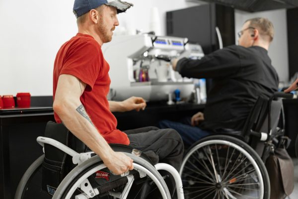 Two men in wheelchairs enjoying time at a coffee shop with an espresso machine in the background.